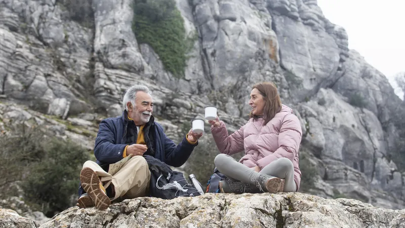 Zwei ältere Wandernde sitzen auf einem Felsen im Gebirge und stoßen mit ihren Bechern an